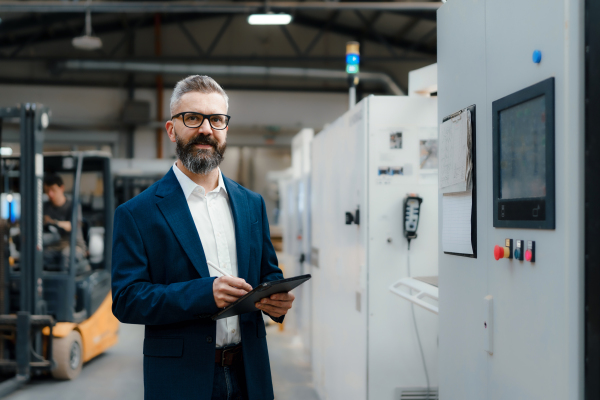 Male project manager standing in modern industrial factory, holding tablet, looking at camera. Manufacturing facility with robotics, robotic arms and automation.