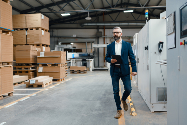 Full length portrait of engineer, project manager walking in modern industrial factory. Manufacturing facility with robotics, robotic arms and automation. Storing products and materials in warehouse.