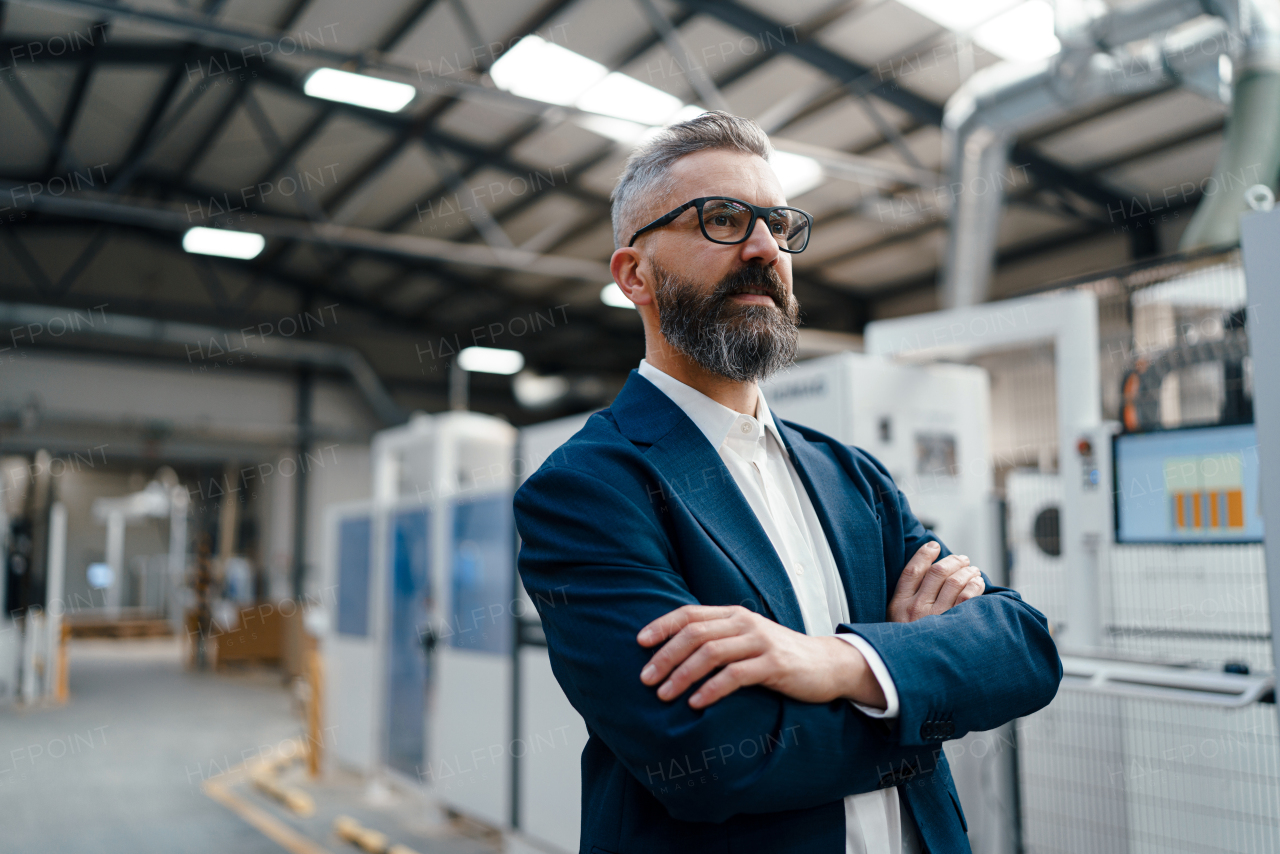 Portrait of male project manager standing in modern industrial factory, low angle with arms crossed. Manufacturing facility with robotics, robotic arms and automation.