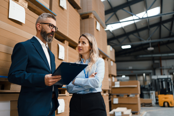 Female warehouse manager talking with logistics employee in warehouse, planning transport of products, goods, talking shipping process. Storing products and materials in warehouse.