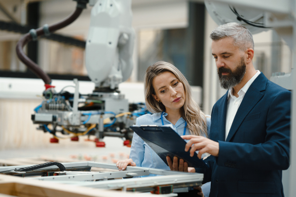 Female engineer and male production manager standing in modern industrial factory, talking about production. Manufacturing facility with robotics, robotic arms and automation.