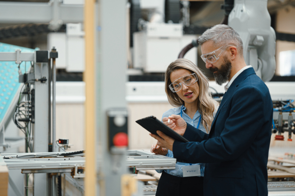 Female engineer and male production manager standing in modern industrial factory, talking about production. Manufacturing facility with robotics, robotic arms and automation.