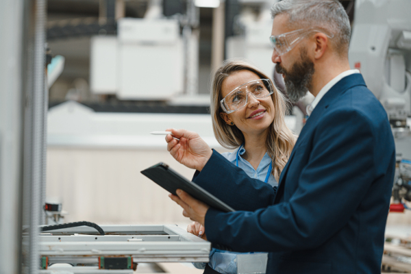 Female engineer and male project manager standing in modern industrial factory, talking about production. Manufacturing facility with robotics, robotic arms and automation.