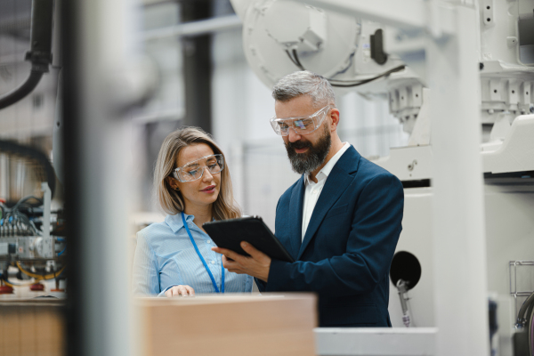 Female engineer and male supervisor standing in modern industrial factory, talking about production. Manufacturing facility with robotics, robotic arms and automation.