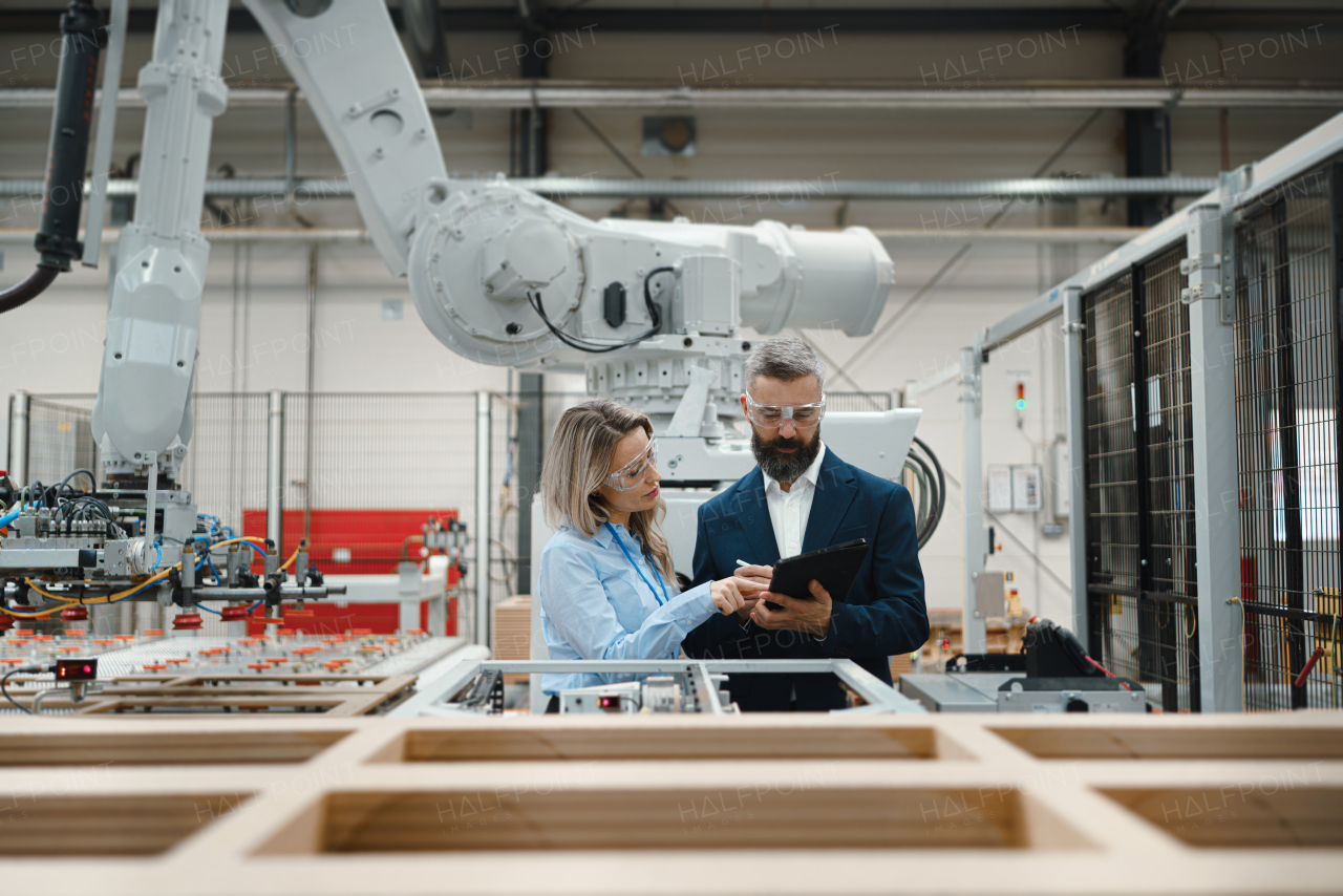 Female engineer and male project manager standing in modern industrial factory, talking about production of wooden furniture. Big furniture manufacturing facility with robotics, robotic arms and automation.