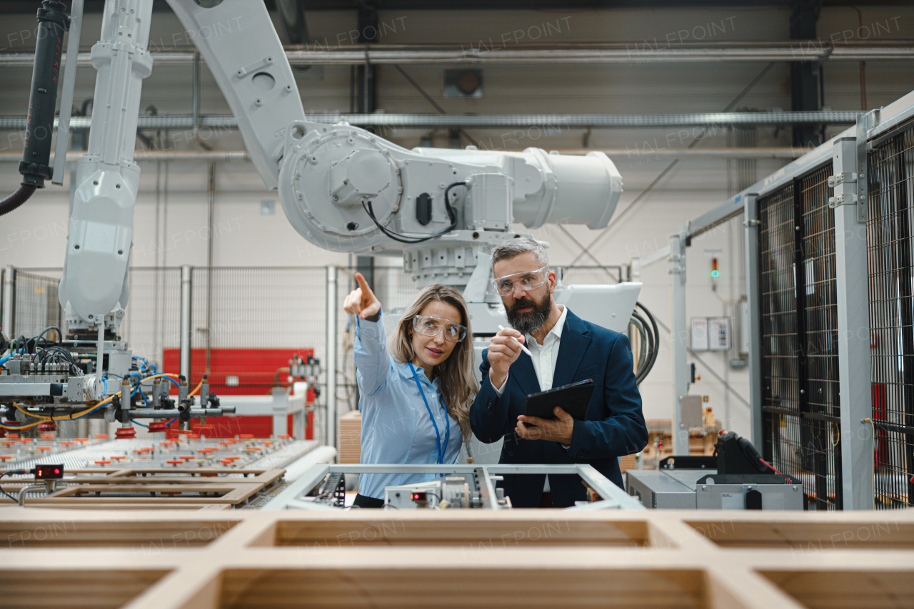 Female engineer and male project manager standing in modern industrial factory, talking about production of wooden furniture. Big furniture manufacturing facility with robotics, robotic arms and automation.