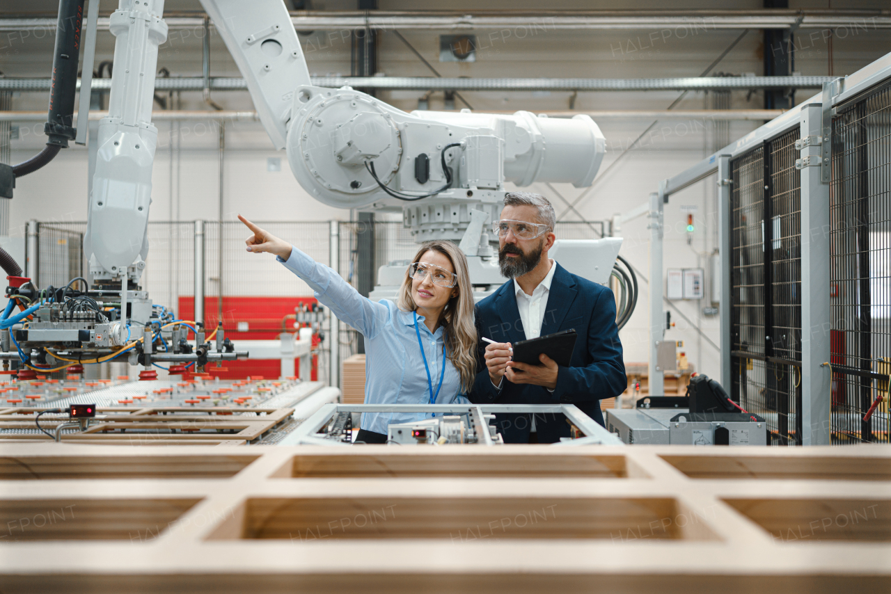 Female engineer and male project manager standing in modern industrial factory, talking about production of wooden furniture. Big furniture manufacturing facility with robotics, robotic arms and automation.