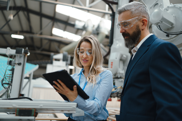 Female engineer and male supervisor standing in modern industrial factory, talking about production. Manufacturing facility with robotics, robotic arms and automation.