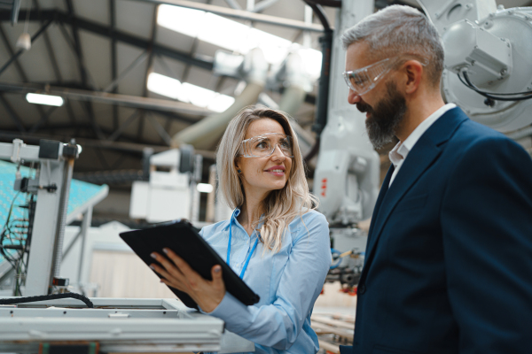 Female engineer and male supervisor standing in modern industrial factory, talking about production. Manufacturing facility with robotics, robotic arms and automation.