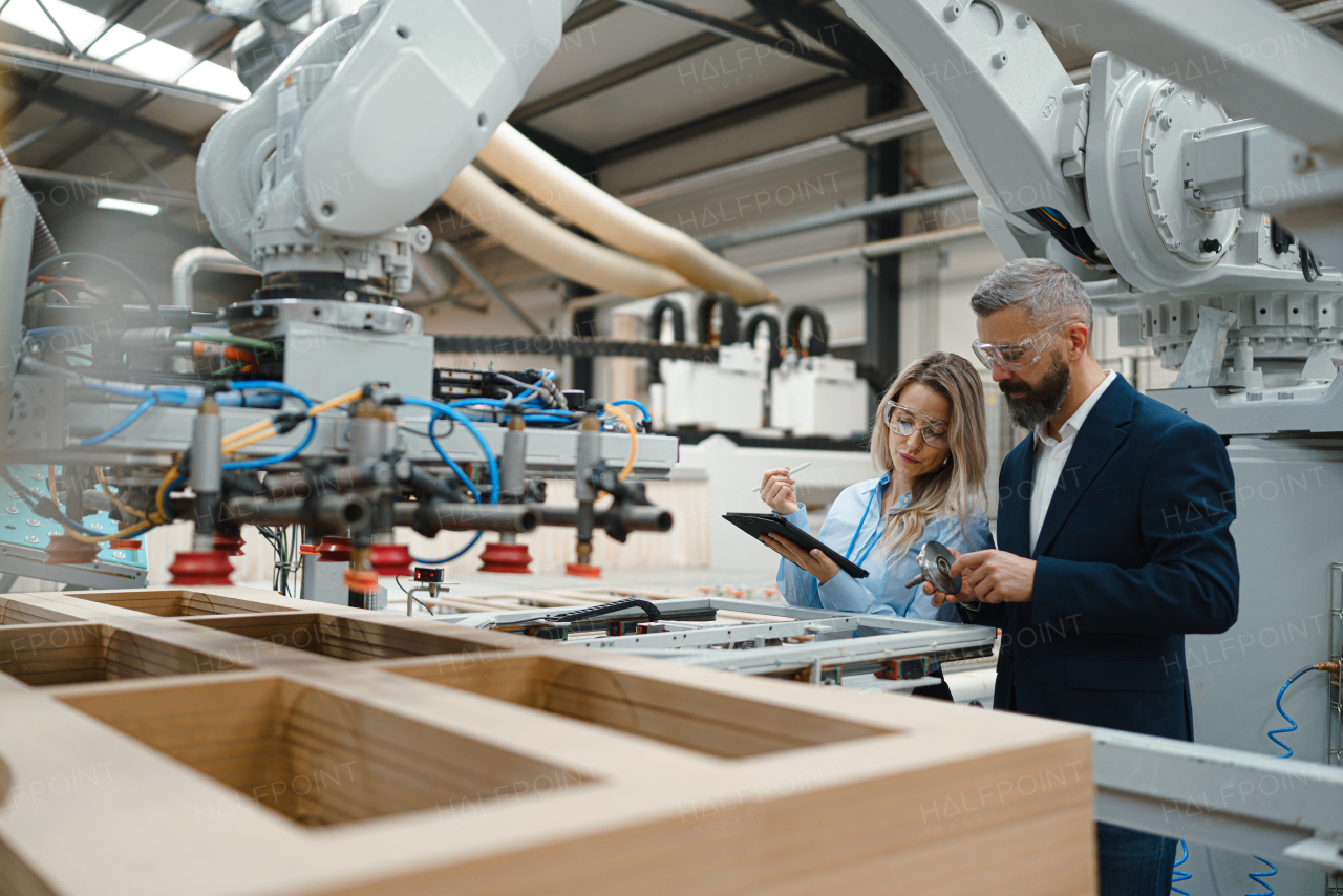 Female engineer and male project manager standing in modern industrial factory, talking about production of wooden furniture. Big furniture manufacturing facility with robotics, robotic arms and automation.