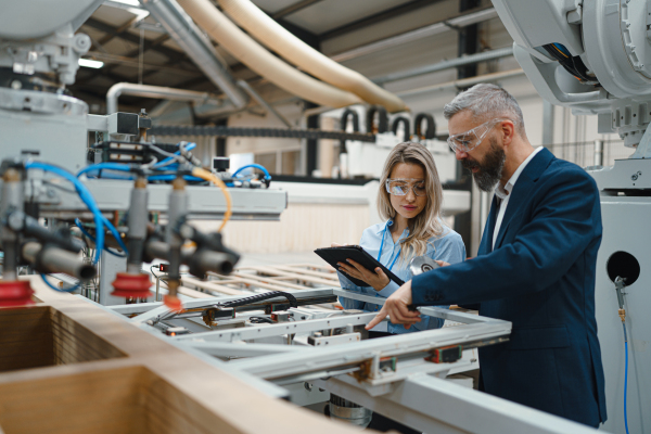 Female engineer and male project manager standing in modern industrial factory, talking about production of wooden furniture. Big furniture manufacturing facility with robotics, robotic arms and automation.