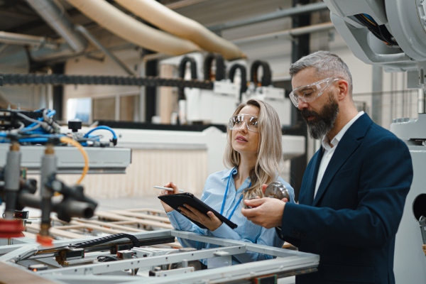 Female engineer and male project manager standing in modern industrial factory by precision robotic arm. Robot assembling wooden furniture in furniture manufacturing facility with industrial robotic manipulators.