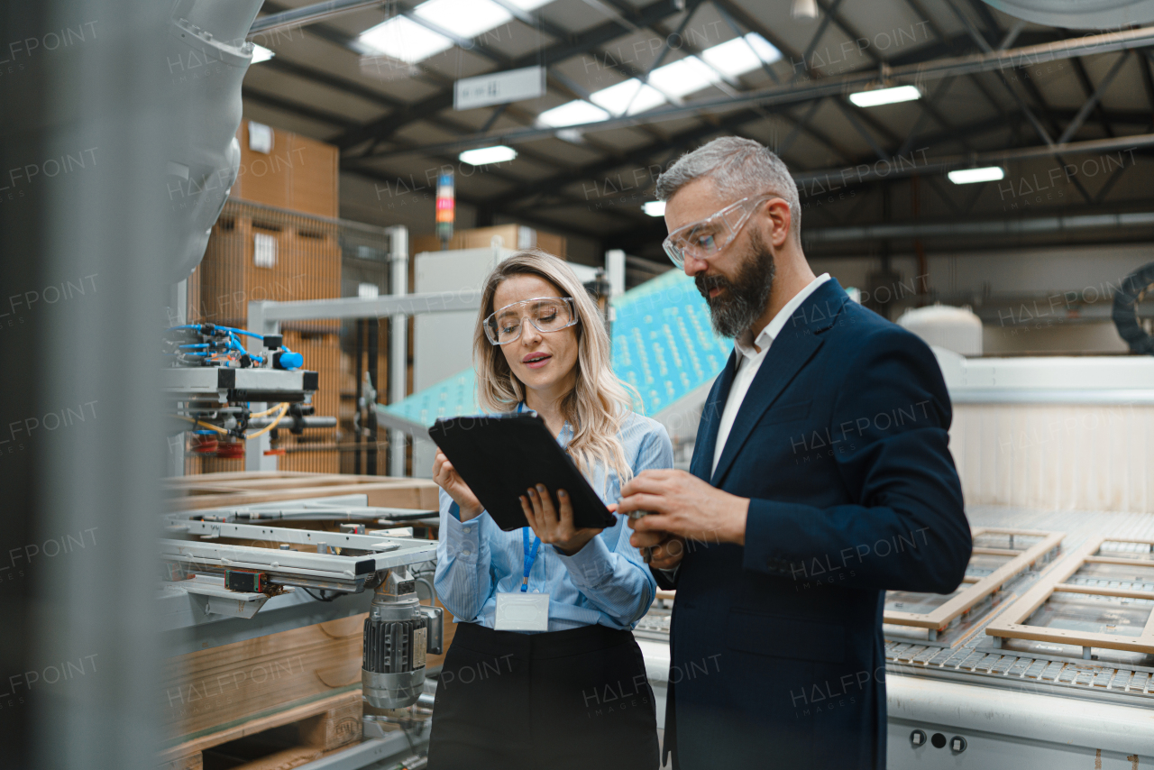 Female engineer and male project manager standing in modern industrial factory, talking about production of wooden furniture. Big furniture manufacturing facility with robotics, robotic arms and automation.