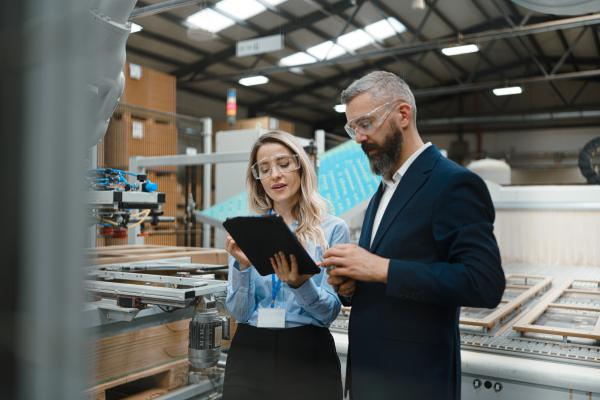 Female engineer and male project manager standing in modern industrial factory, talking about production of wooden furniture. Big furniture manufacturing facility with robotics, robotic arms and automation.