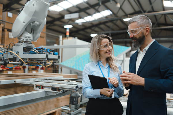 Female engineer and male project manager standing in modern industrial factory, talking about production of wooden furniture. Big furniture manufacturing facility with robotics, robotic arms and automation.