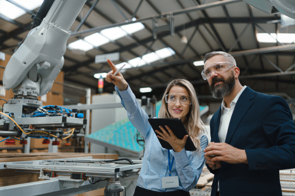 Female engineer and male project manager standing in modern industrial factory by precision robotic arm. Robot assembling wooden furniture in furniture manufacturing facility with industrial robotic manipulators.