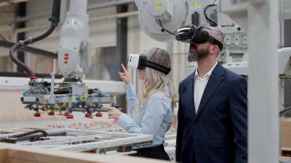 Female engineer and male project manager standing in modern industrial factory by precision robotic arm, using VR. Robot assembling wooden furniture in furniture manufacturing facility with industrial robotic manipulators. Virtual reality in manufacturing industry.