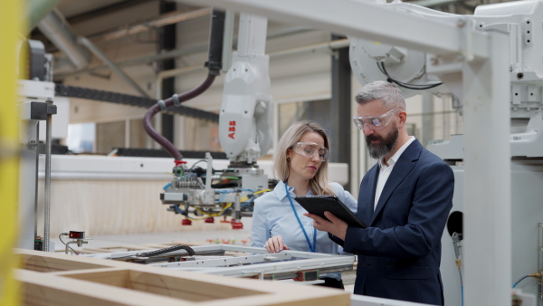 Female engineer and male project manager standing in modern industrial factory, talking about production of wooden furniture. Big furniture manufacturing facility with robotics, robotic arms and automation.