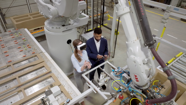 Female engineer and male project manager standing in modern industrial factory by precision robotic arm, using VR. Robot assembling wooden furniture in furniture manufacturing facility with industrial robotic manipulators. Virtual reality in manufacturing industry.