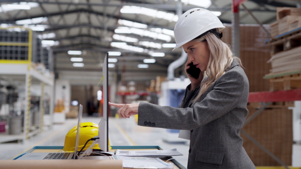 Female engineer, company director, manager in modern industrial factory making call on smartphone. Production control station with pc, computer in manufactury.