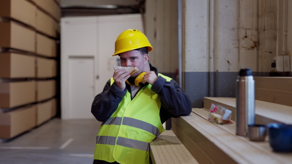 Worker with Down syndrome having break in industrial factory, warehouse, eating sandwich. Concept of workers with disabilities, support in workplace.