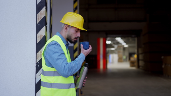 Video of worker taking break from work, drinking coffee, standing on loading dock, outdoors. Employee drinking warm tea from thermor.