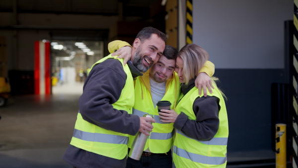 Man with Down syndrome and his colleagues taking break from work, drinking coffee, hot tea from thermos, standing on loading dock. Concept of workers with disabilities, support in workplace.