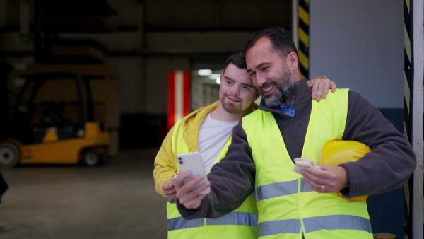 Man with Down syndrome and his colleague taking break from work, taking selfie on smartphone and having fun. Concept of workers with disabilities, support in workplace.