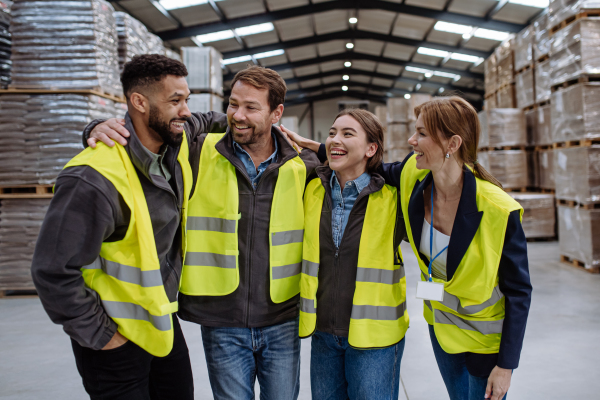 Full team of warehouse employees laughing in warehouse, holding each other by shoulders. Team of workers in reflective clothing in modern industrial factory, heavy industry, manufactrury. Group portrait.