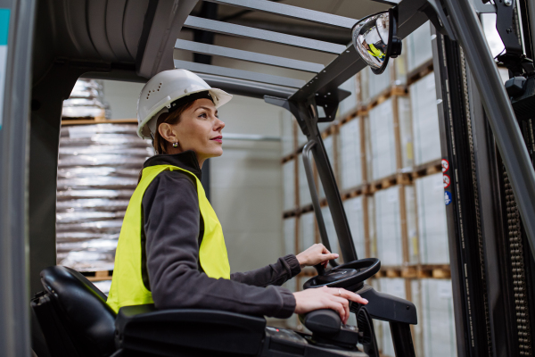 Portrait of female warehouse worker driving forklift. Warehouse worker preparing products for shipmennt, delivery, checking stock in warehouse.