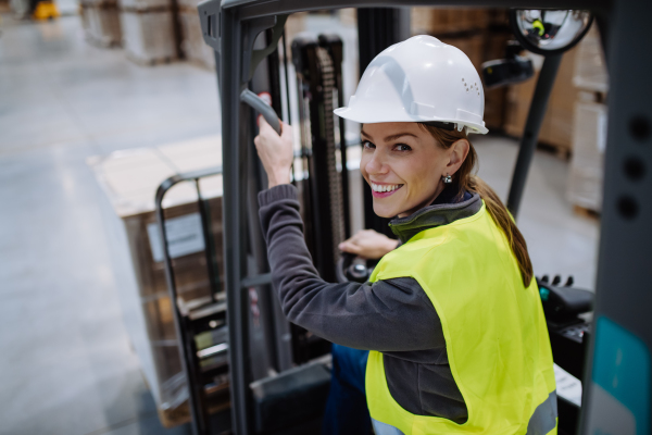 Portrait of female warehouse worker driving forklift. Warehouse worker preparing products for shipmennt, delivery, checking stock in warehouse.