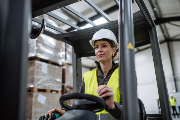 Portrait of female warehouse worker driving forklift. Warehouse worker preparing products for shipmennt, delivery, checking stock in warehouse.