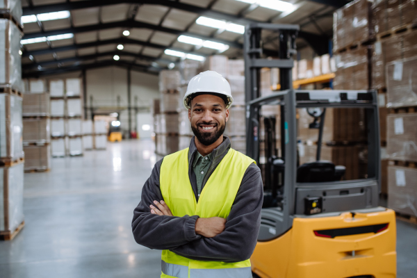 Portrait of multiracial warehouse worker standing by forklift. Warehouse worker preparing products for shipmennt, delivery, checking stock in warehouse. Banner with copy space.