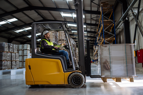 Side view of forklift in warehouse with male driver. Warehouse worker preparing products for shipmennt, delivery, checking stock in the warehouse.