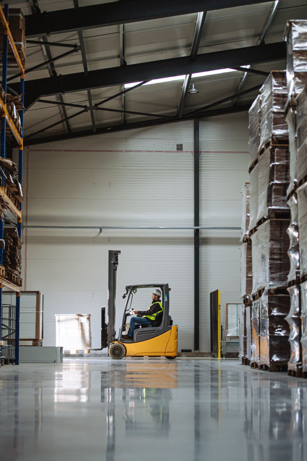 Side view of forklift in warehouse with male driver. Warehouse worker preparing products for shipmennt, delivery, checking stock in the warehouse.