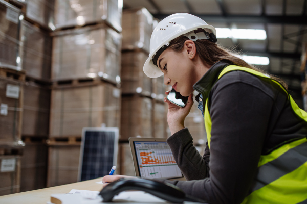 Female warehouse worker checking delivery, stock in warehouse on company pc while phone calling with contractor. Warehouse manager using warehouse management software, app.