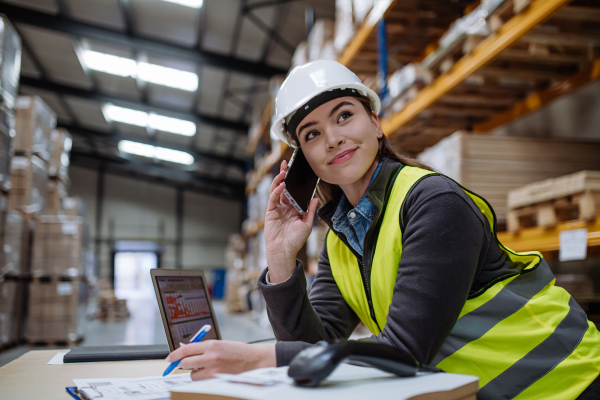 Female warehouse worker checking delivery, stock in warehouse on company pc while phone calling with contractor. Warehouse manager using warehouse management software, app.