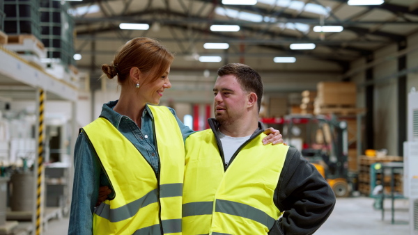 Young man with Down syndrome working in warehouse, colleague talking with him. Concept of the workers with disabilities, support in workplace.