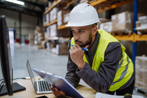 Portrait of warehouseman checking delivery, stock in warehouse on company pc, computer. Warehouse manager using warehouse management software, app.