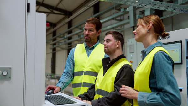 Young man with Down syndrome working in warehouse, colleague teaching him how to work with computer. Concept of workers with disabilities, support in workplace.