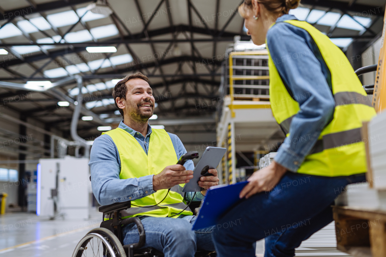 Man in wheelchair working in modern industrial factory, talking with female coworker. Concept of workers with disabilities, accessible workplace for employees with mobility impairment.