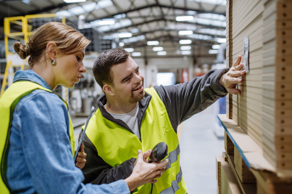 Young man with Down syndrome working in warehouse, female colleague teaching him how to scan products with scanner. Concept of workers with disabilities, support in workplace.