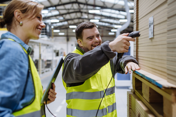 Young man with Down syndrome working in warehouse, female colleague teaching him how to scan products with scanner. Concept of workers with disabilities, support in workplace.