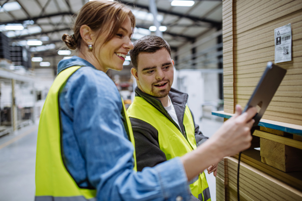 Young man with Down syndrome working in warehouse, female colleague teaching him how to scan products with scanner. Concept of workers with disabilities, support in workplace.