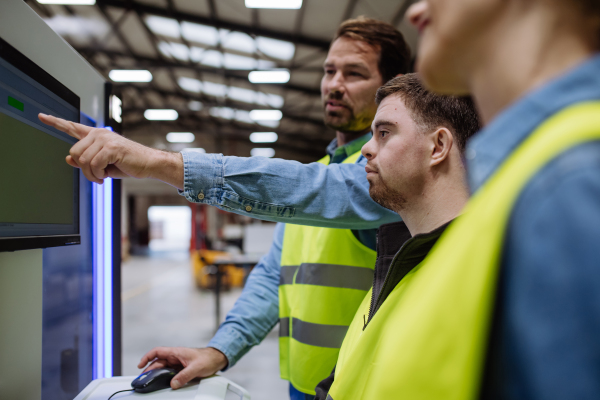 Young man with Down syndrome working in warehouse, colleague teaching him how to work with computer. Concept of workers with disabilities, support in workplace.