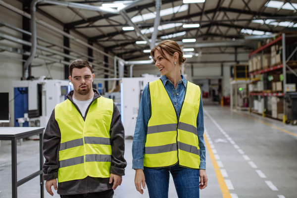 Young man with Down syndrome working in warehouse, colleague talking with him. Concept of the workers with disabilities, support in workplace.