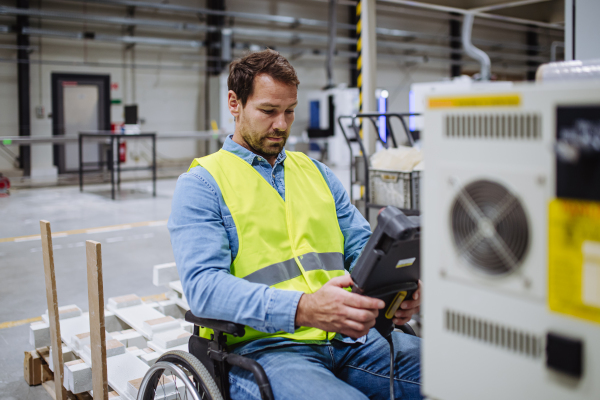 Man in wheelchair working in modern industrial factory, scanning barcodes with scanner. Concept of workers with disabilities, accessible workplace for employees with mobility impairment.