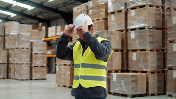 Video of male warehouse worker standing in warehouse. Warehouse employee putting on a working helmet.