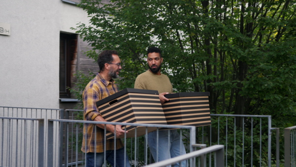 Man helping his friend move into new apartment in building. Man is moving into new flat, carrying box from the moving truck and phone calling.