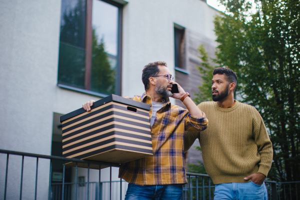 Man helping his friend move into new apartment in building. Man is moving into new flat, carrying box from the moving truck and phone calling.